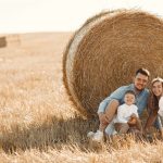 Smiling family sitting together by a hay bale in a sunny rural field, capturing a joyful moment.