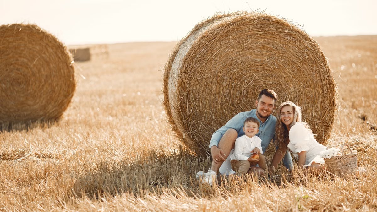 Smiling family sitting together by a hay bale in a sunny rural field, capturing a joyful moment.