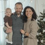 A joyful family with a baby poses next to a decorated Christmas tree indoors.