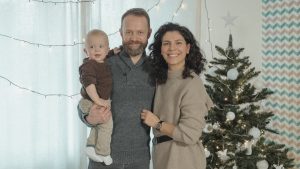 A joyful family with a baby poses next to a decorated Christmas tree indoors.